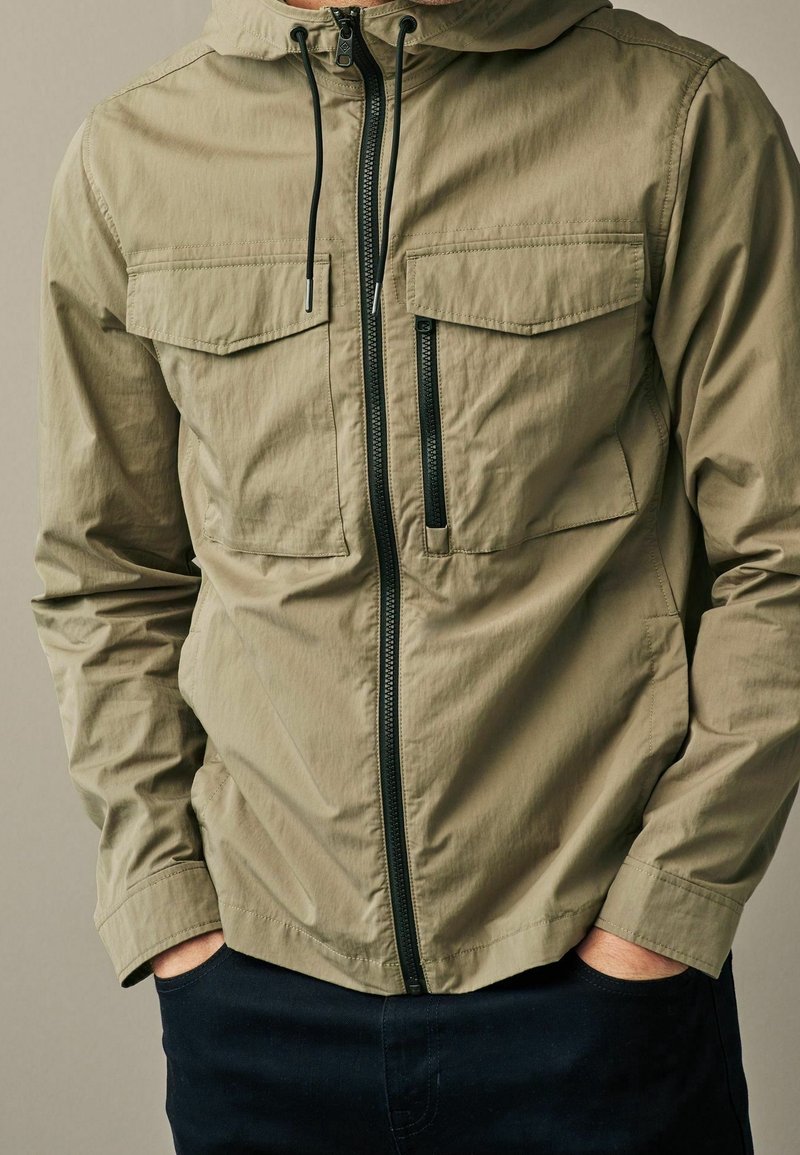 Veste légère couleur olive, avec fermeture éclair à l'avant, capuche réglable et deux poches poitrine. Elle présente une texture lisse et des accents noirs.