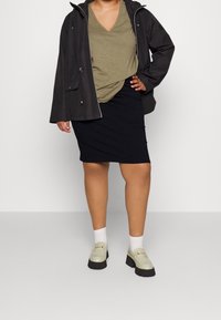Persona vestida con una camiseta verde oliva de pico, chaqueta negra, falda negra hasta la rodilla, calcetines blancos y mocasines de plataforma beige, de pie con la mano en la cadera.