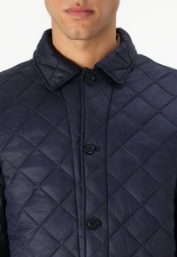 Jachetă din nylon matlasat de culoare navy, cu un guler clasic, patru butoane negre și un model de cusătură în formă de diamant, creată pentru un fit structurat.