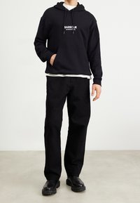 Černá mikina s kapucí s logem "BARBOUR INTERNATIONAL", klokaní kapsa, v kombinaci s černými volnými kalhotami a robustními černými botami.
