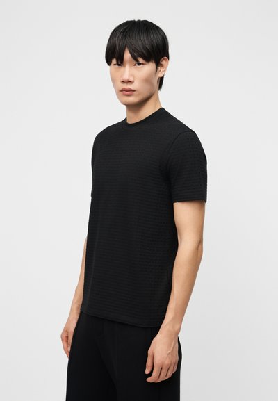 Νεαρός άνδρας με κοντά μαύρα μαλλιά, φοράει μαύρο κοντομάνικο πουκάμισο με υφή, στέκεται μπροστά σε μια απλή ανοιχτογκρι φόντο.