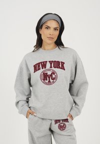 Felpa grigia con stampa rossa "NEW YORK" sul davanti, caratterizzata da un logo rotondo. Tessuto morbido, vestibilità comoda, abbinata a pantaloni della tuta coordinati.
