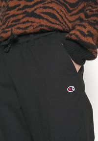 Black pants with a textured design, drawstring waist, and side pockets. Features a small embroidered logo on the left side.