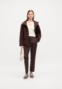Brown wool jacket with hood, cream knit top, brown trousers, and gray sneakers. Features a small beige crossbody bag.