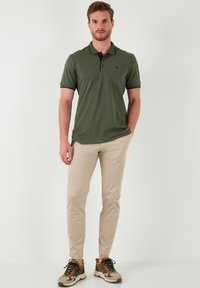 Olívazöld póló fekete gallér részletekkel, bézs slim-fit nadrág, és színes, fűzős sneakers.