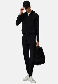 Svart zip-up hoodie, matchande svarta smala byxor, vita sneakers och svart keps, med en stilren svart ryggsäck i handen.