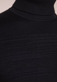 Pull en col roulé noir en tricot côtelé, présentant des rayures texturées horizontales au niveau de la poitrine.