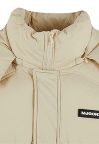 Beige padded jacket with a high collar, front zipper, and black logo patch. Features subtle stitching and smooth texture.