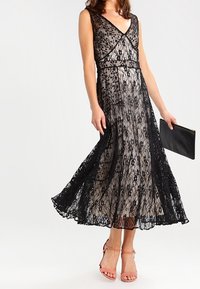 Svart spetsklänning med V-ringning, ärmlös design, lager ovanpå en nude-underklänning, hel längdkjol och detaljerad blomstermönster.