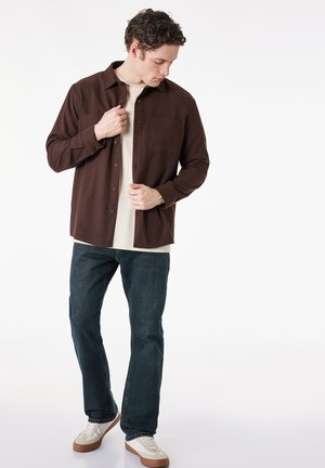 Mann knöpft ein dunkelbraunes Hemd über einem beigen T-Shirt zu, trägt blaue Jeans und weiße Sneaker und steht vor weißem Hintergrund.