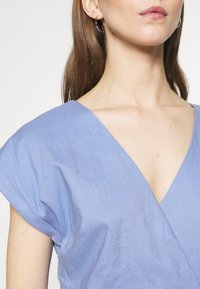 Femme portant un haut bleu clair à manches courtes en V, avec des créoles, montrant le cou et la zone des épaules sur fond blanc.