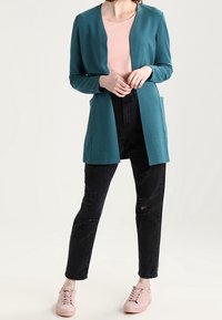 Tyrkysový strukturovaný cardigan s otevřeným přední částí a dvěma kapsami, nošený přes světle růžové tričko, v kombinaci s černými roztrhanými džíny a světle růžovými teniskami.