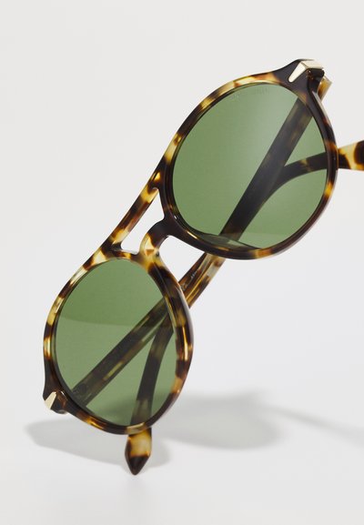 Ochelari de soare din tortoise brown cu lentile verzi, forma rotundă și brațe elegante cu un accent subtil aproape de ramă.