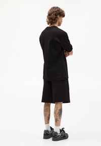 Sort bomulds top med korte ærmer og matchende shorts, der har en afslappet pasform. Grå sneakers og hvide sokker fuldender outfittet. Tatoveringer er synlige.