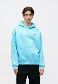 Tânăr îmbrăcat cu un hanorac bleu deschis cu un logo Jumpman negru și pantaloni negri, stând în fața unui fundal alb simplu.