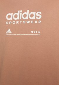 Světle hnědé bavlněné tričko s výrazným bílým textem "adidas SPORTSWEAR" a logem značky, s dalšími tištěnými motivačními frázemi níže.