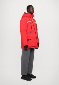 Rote gepolsterte Jacke mit Kapuze, reflektierenden Streifen, mehreren Taschen und schwarzen Akzenten. Kombiniert mit grauen weiten Hosen und schwarzen Schuhen.