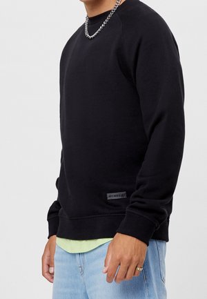 Sudadera negra con cuello redondo, puños acanalados, tela texturizada y un parche con logo en la parte inferior. Se lleva sobre una camisa verde claro.