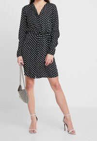 Svart klänning med vita prickar, långärmad och med åtsittande midja, kombinerad med silverfärgade klackar med remmar och en liten kedjeväska.