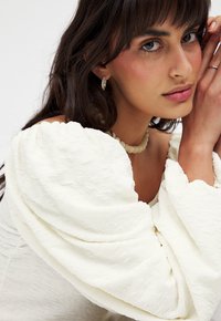 Witte textuurstof top met pofmouwen en gerimpelde details. Model draagt een bewerkte ketting en zilveren oorbellen.