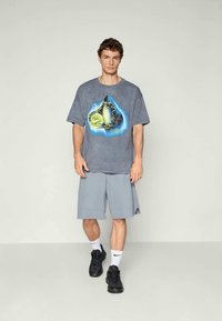 Ung mand iført en grå grafisk t-shirt, lysegrå knælange shorts, hvide Nike-sokker og sorte sneakers står foran en ensfarvet baggrund.