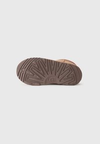 Suela de goma marrón con un patrón texturizado en zigzag y el logo de UGG. Parte superior en color crema con dos correas, que ofrece un diseño minimalista.