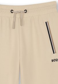 Pantalon de survêtement beige avec taille à cordon, détail rayé noir et beige sur la poche latérale, et logo partiel "BOSS" sur la cuisse.