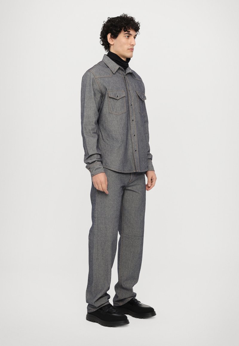 Grå denimskjorta med bröstfickor, matchande avsmalnade grå byxor och svarta skor. Outfiten har kontrasterande sömmar och en avslappnad passform.