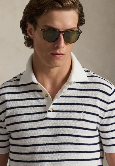 Striped polo shirt in white with navy stripes, featuring a classic collar and two gold buttons, worn with tortoiseshell sunglasses.