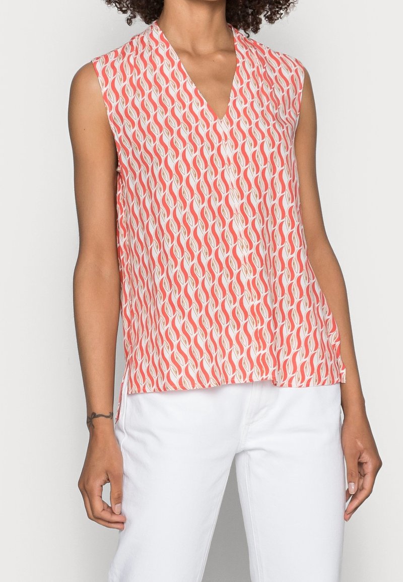 Personne portant un chemisier sans manches à encolure en V avec un motif géométrique ondulé rouge et blanc et un pantalon blanc, debout devant un fond uni.