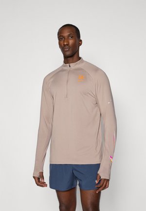 Šviesiai rudos spalvos ilgomis rankovėmis sportinė palaidinė su ketvirtiniu užtrauktuku, puošta oranžiniu logotipu ir daugiaspalviais akcentais ant rankovių, derinama su tamsiai mėlynomis šortais.