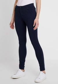 Mørkeblå strækleggings med en glat tekstur og slim fit, der har en frontlomme med lynlås og knaplukning. Bæres med hvide sneakers.