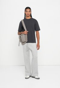 Νεαρός άνδρας με μακριές πλεξίδες φοράει μαύρο t-shirt, ανοιχτό γκρι φόρμα, λευκά αθλητικά παπούτσια και κρατά μια γκρι τσάντα ώμου σε λευκό φόντο.