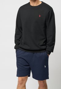 Hombre con sudadera negra de manga larga y pantalones cortos azules marino, con las manos en los bolsillos, llevando logos bordados de jugador de polo visibles en ambas prendas.