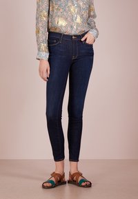 Vaqueros ajustados azul oscuro con cintura alta, combinados con una camisa de botones de estampado floral y sandalias multicolores. Textura de mezclilla suave.