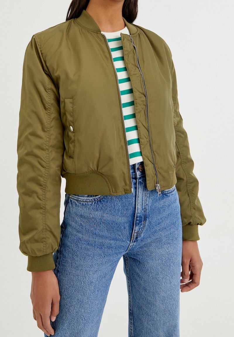 Woman wearing olive green bomber jacket over green and white striped shirt with light blue high-waisted jeans, standing with arms relaxed.