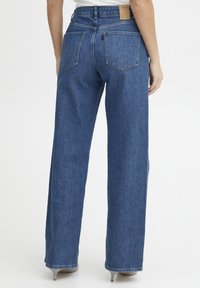 Jeans de tiro alto en azul con piernas anchas, diseño de cinco bolsillos y costuras visibles; tela de mezclilla texturada; parche de cuero en la cintura.