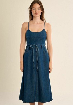 Donna che indossa un abito midi in denim blu senza maniche con spalline sottili e cintura annodata in vita, in piedi davanti a uno sfondo beige uniforme.