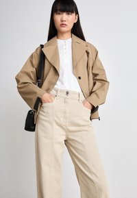 Tann oversized jakke med svarte knapper, hvit skjorte med krage, og vide beige bukser. Svart crossover-veske gir kontrast og funksjonalitet.