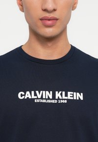 Jūras spēku zila kokvilnas T-kreklu ar izteiktu baltu "CALVIN KLEIN" tekstu, ar "ESTABLISHED 1968" zem tā. Standarta apaļa kakla izgriezums un īsas piedurknes.