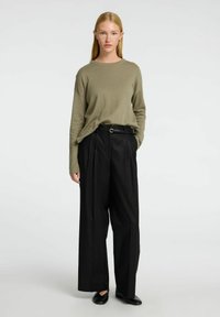 Olivgrüner Strickpullover mit lockerer Passform, kombiniert mit hochgeschnittener, schwarzer weit geschnittener Hose und einfachen schwarzen Slippern.