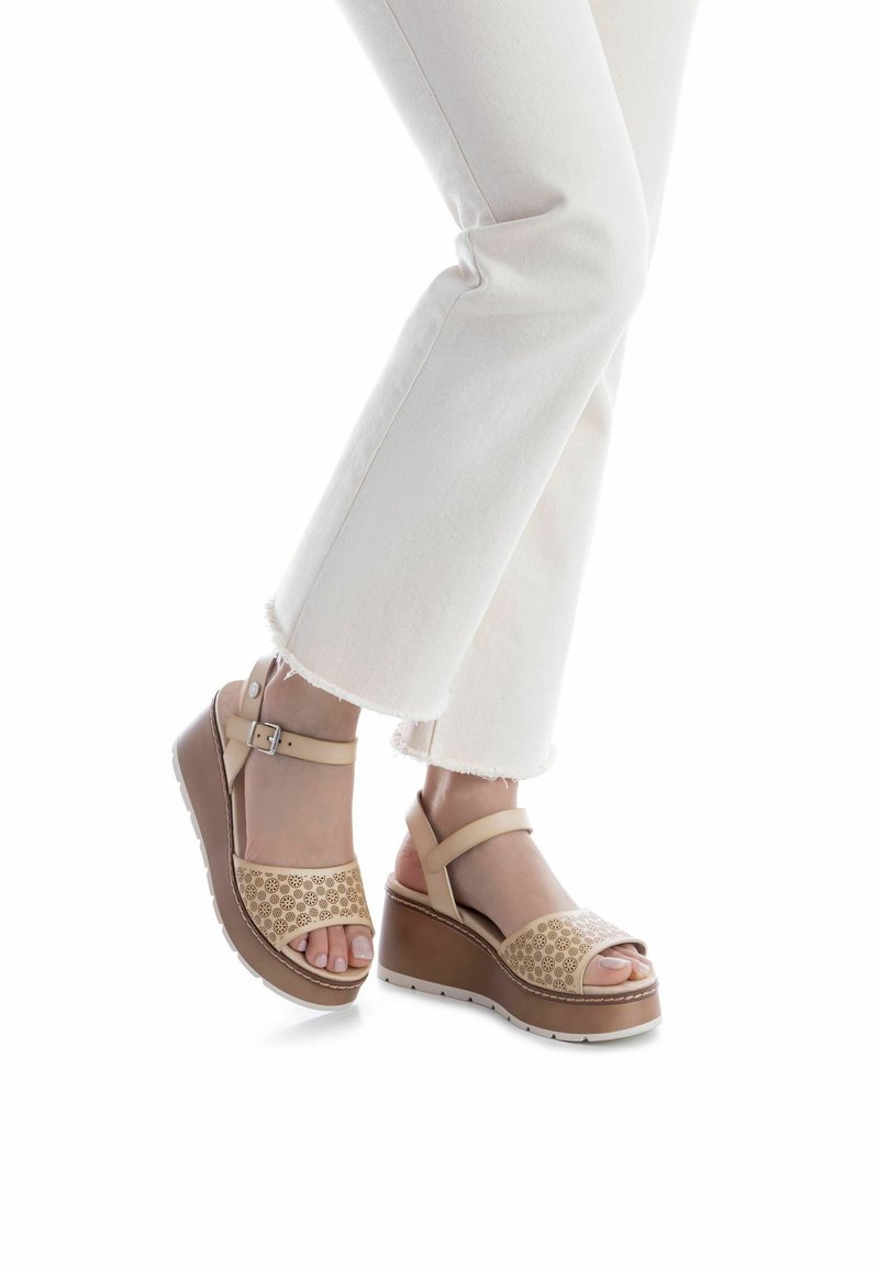 Sandalias de cuña beige con patrón floral recortado, correa ajustable en el tobillo y suela marrón claro. Llevadas con vaqueros blancos de dobladillo deshilachado.