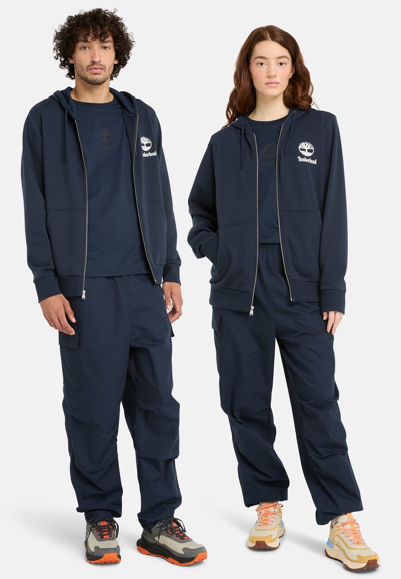 Sudadera con cremallera en color azul marino con cordones, combinada con pantalones cargo azul marino y calzado en contraste con acentos en gris, naranja y beige.