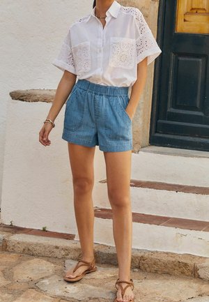 Chemise blanche avec des détails en dentelle à œillets, deux poches avant. Shorts en denim avec une taille élastique et des poches latérales. Sandales beiges.