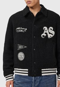 Sort tekstureret varsity-jacket med hvide broderede lapper, stribede manchetter og kant, båret af en person med blå jeans.