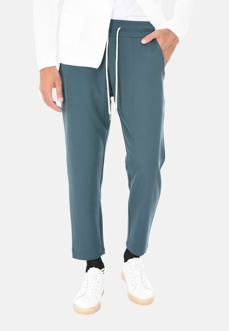 Uomo che indossa pantaloni teal con cordino, calze nere, scarpe da ginnastica bianche e una giacca bianca con texture, con una mano in tasca.