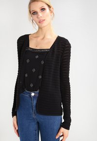 Svart åpen cardigan med teksturert stoff, lange ermer og ribbestrikkede detaljer. Båret over en svart topp med geometriske mønstre og blå jeans.