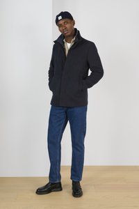 Giacca di pile navy con colletto alto, chiusura a bottoni, abbinata a una camicia chiara e jeans in denim blu. Mocassini neri completano il look.