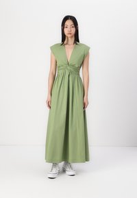 Abito lungo verde con scollatura a V profonda e maniche a cappuccio. Realizzato in un tessuto morbido con dettaglio di vita torcigliato, abbinato a sneakers bianche.