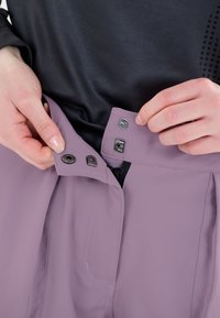 Lila byxor med en mjuk yta, med tryckknapp och dragkedja. Midjebandet har synliga sömmar, delvis synligt genom händer.